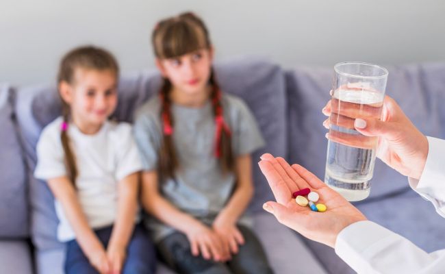 Zdjęcie przedstawia osobę dorosłą wręczającą dzieciom tabletki.
